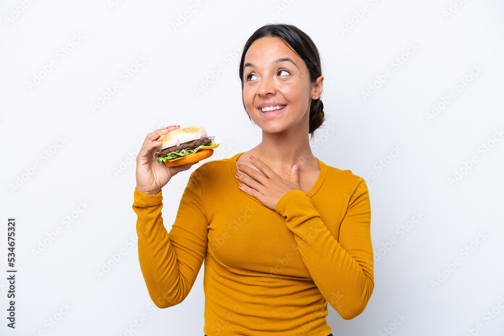 Young hispanic woman holding a burger isolated on white background looking up while smiling Young hispanic woman holding a burger isolated on white background looking up while smiling