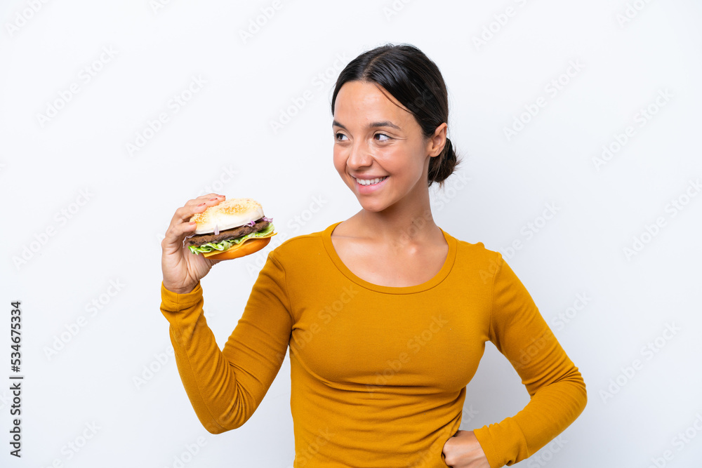Young hispanic woman holding a burger isolated on white background looking to the side and smiling Young hispanic woman holding a burger isolated on white background looking to the side and smiling