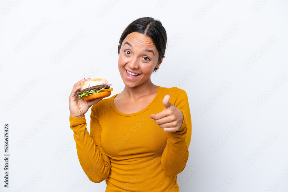 Young hispanic woman holding a burger isolated on white background with thumbs up because something good has happened Young hispanic woman holding a burger isolated on white background with thumbs up because something good has happened