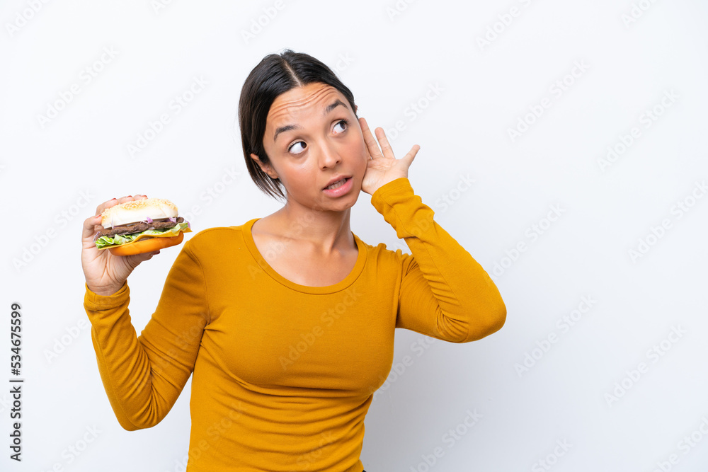 Young hispanic woman holding a burger isolated on white background listening to something by putting hand on the ear Young hispanic woman holding a burger isolated on white background listening to something by putting hand on the ear