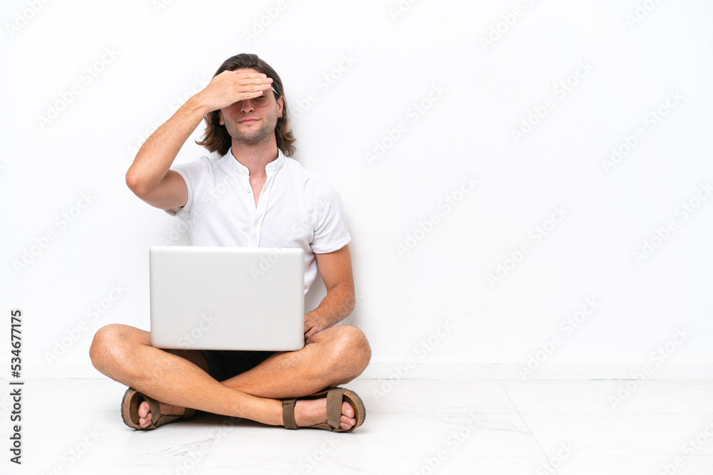 Young handsome man with a laptop sitting on the floor isolated on white background covering eyes by hands. Do not want to see something Young handsome man with a laptop sitting on the floor isolated on white background covering eyes by hands. Do not want to see something