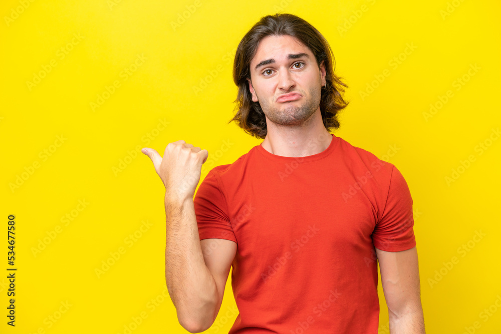 Caucasian handsome man isolated on yellow background unhappy and pointing to the side Caucasian handsome man isolated on yellow background unhappy and pointing to the side