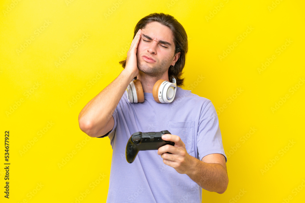 Young handsome caucasian man playing with a video game controller over isolated on yellow background with headache Young handsome caucasian man playing with a video game controller over isolated on yellow background with headache