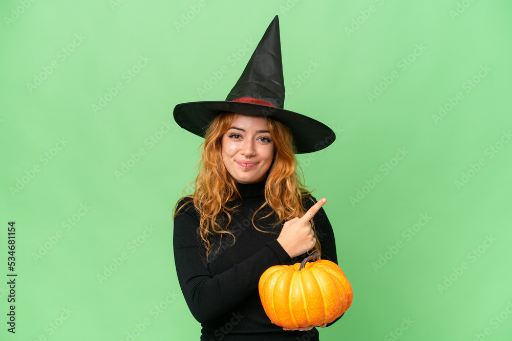 Young caucasian woman costume as witch holding a pumpkin isolated on green screen chroma key background pointing to the side to present a product Young caucasian woman costume as witch holding a pumpkin isolated on green screen chroma key background pointing to the side to present a product