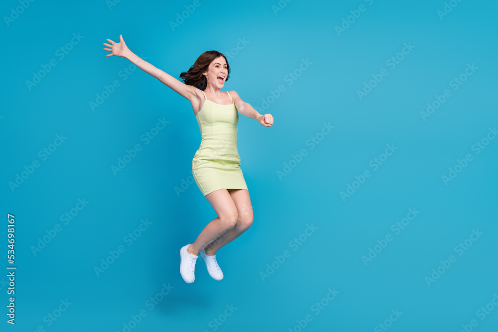 Full length photo of excited funky lady wear green outfit jumping high arms sides looking empty space isolated blue color background