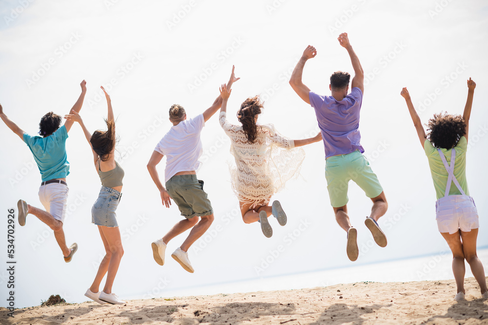 Full size rear behind portrait of company crazy ecstatic fellows jumping raise fists sunny weather water sand beach outdoors Full size rear behind portrait of company crazy ecstatic fellows jumping raise fists sunny weather water sand beach outdoors