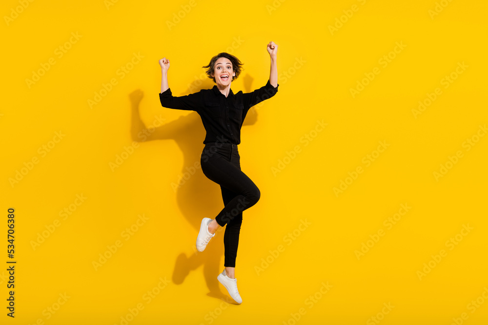 Photo of charming lucky girl dressed formal shirt jumping high rising fists empty space isolated yellow color background