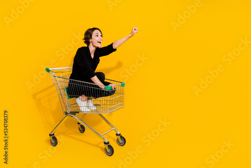 Photo of sweet charming girl dressed formal shirt riding shopping tray rising fist empty space isolated yellow color background