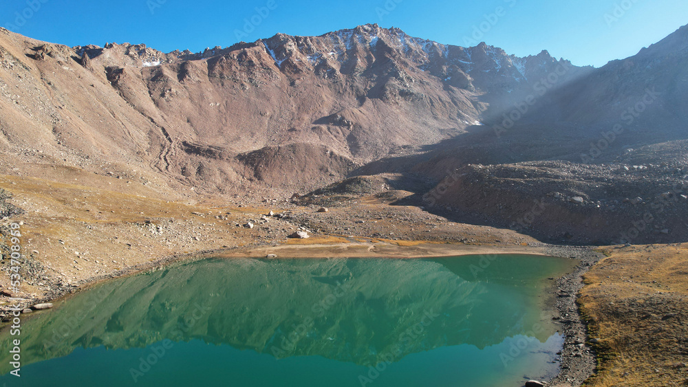 The emerald color of the water of a mountain lake. Drone view of a green mountain lake with a reflection of the mountains. Smooth transparent water. There are large stones nearby. Dry grass Kazakhstan The emerald color of the water of a mountain lake. Drone view of a green mountain lake with a reflection of the mountains. Smooth transparent water. There are large stones nearby. Dry grass Kazakhstan