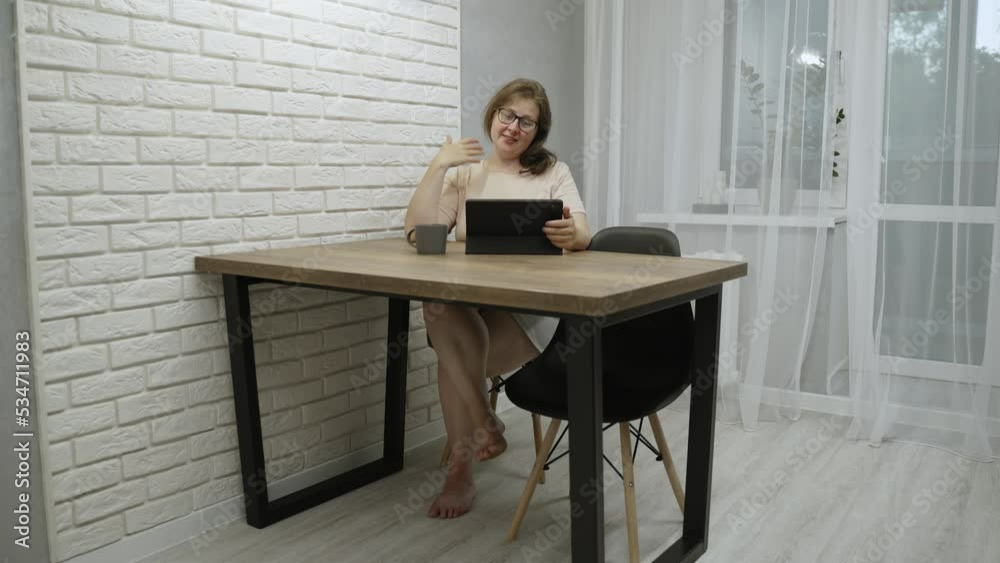 Overweight woman with glasses makes video call on tablet in apartment. Overweight woman with glasses makes video call on tablet in apartment.