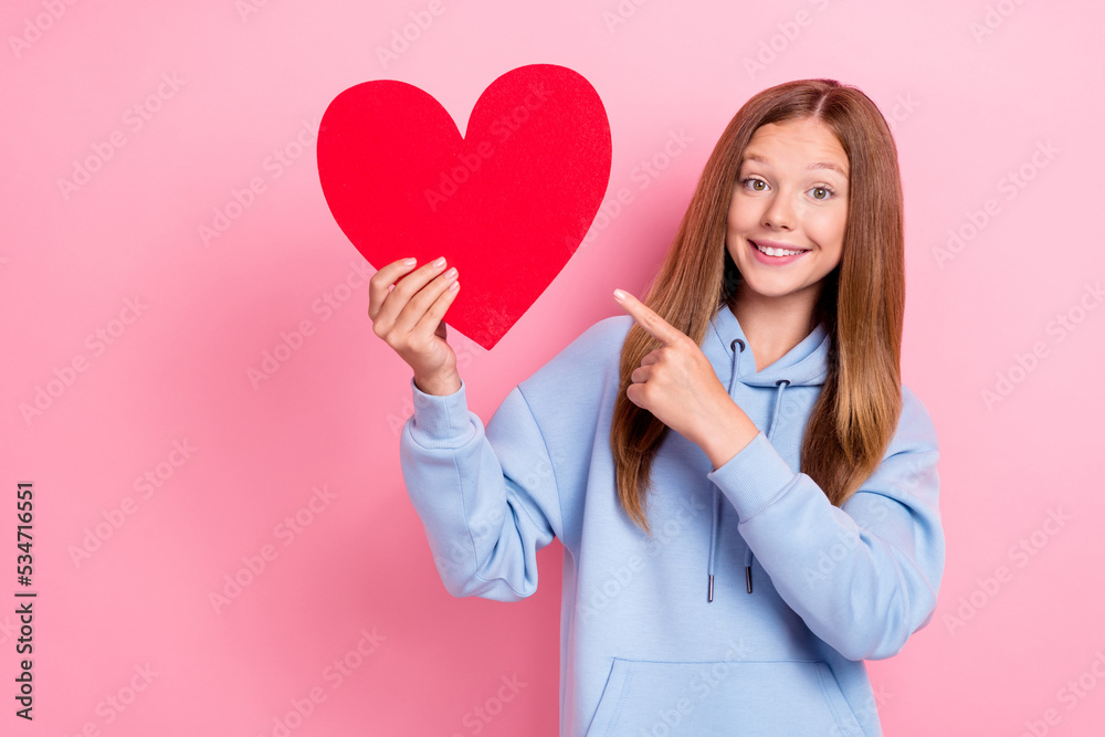 Photo of young little cute pretty excited positive schoolgirl wear stylish hoodie finger point hold red paper heart like isolated on pink color background