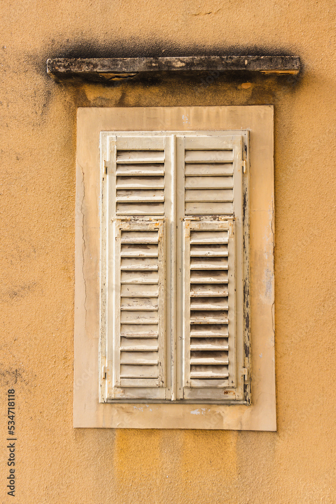 Closed window with white shutters on the yellow wall of an old house