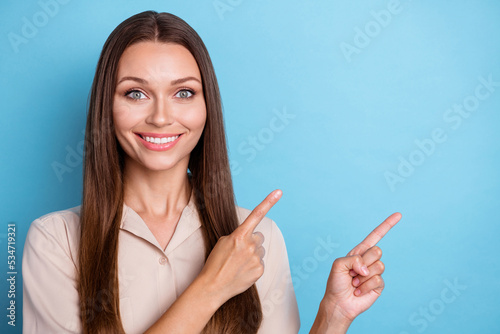 Photo of adorable gorgeous cheerful girl with long hairstyle dressed beige blouse directing empty space isolated on blue color background