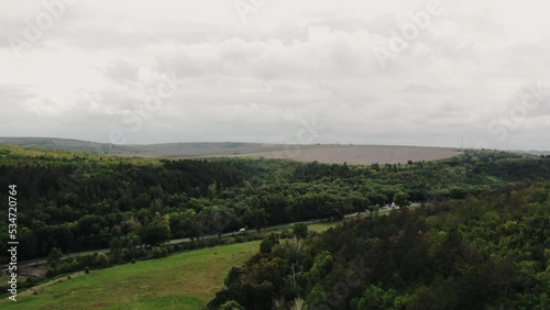 Cinematic aerial top view of the road with traffic between green and yellow trees on the autumn field near the windy forest. Drone shot of a road in the forest and autumn cloudy sky.