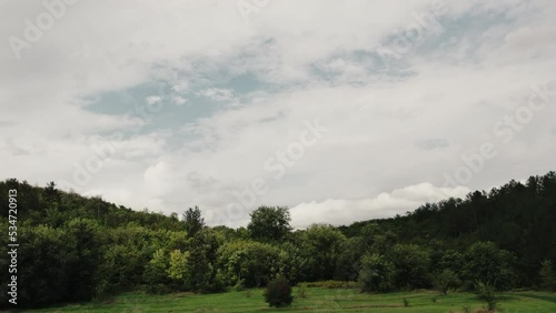 Cinematic aerial top view of the small field between green and yellow trees near the windy forest. Drone shot of a road in the forest and autumn cloudy sky.