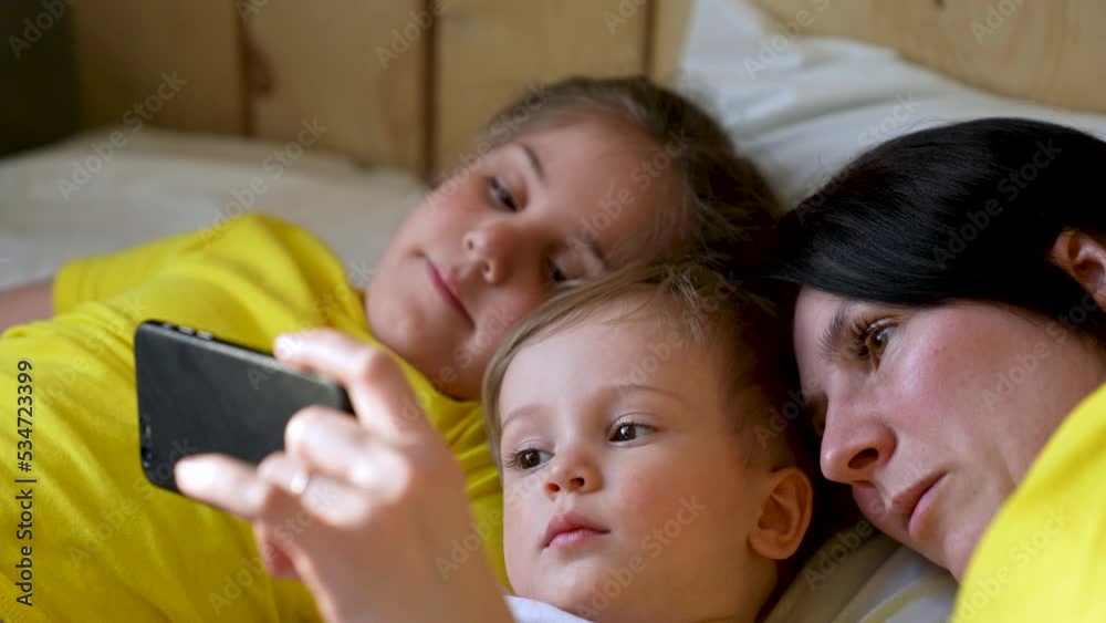Happy family concept. Mom, daughter and son are play on bed. Child watching video on smartphone. Family leisure at home. Boy and girl with mom are watching movie on smartphone. Home education concept Happy family concept. Mom, daughter and son are play on bed. Child watching video on smartphone. Family leisure at home. Boy and girl with mom are watching movie on smartphone. Home education concept