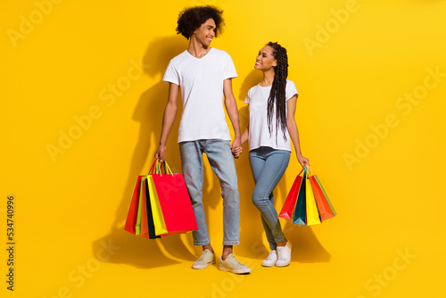 Full length portrait of two positive lovely people hold hands boutique packages isolated on yellow color background