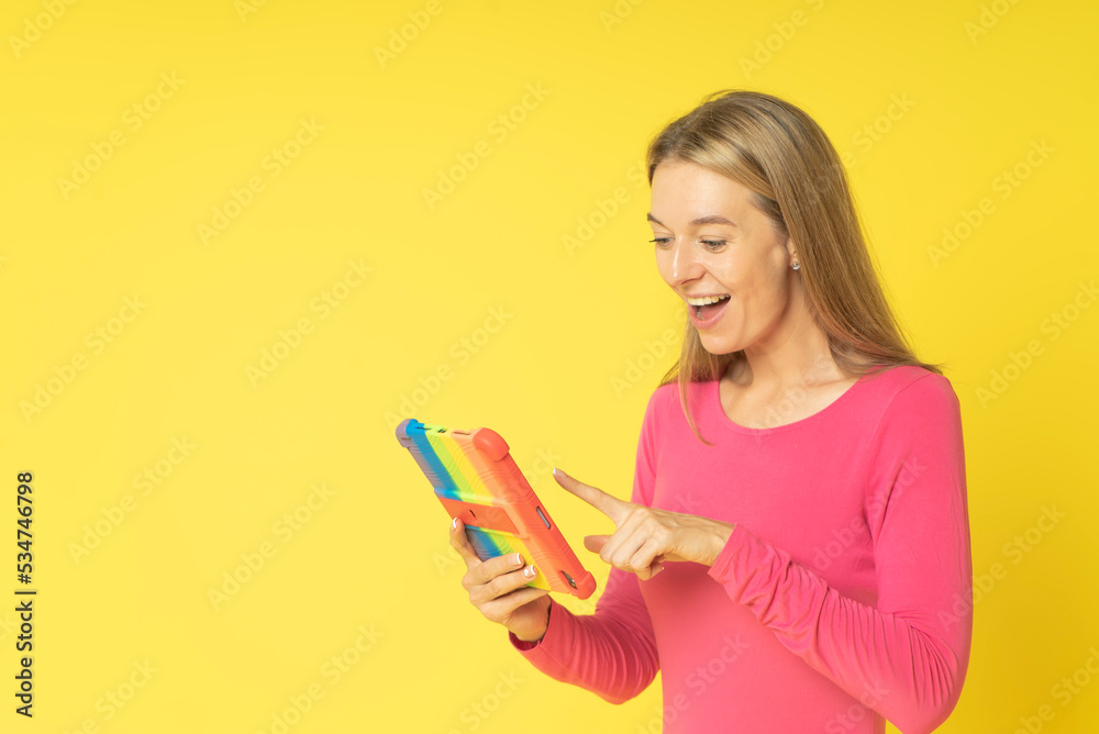 Beautiful excited smiling woman in red dress reading a text message or look browse tablet in rainbow cover app button download isolated over yellow color background Beautiful excited smiling woman in red dress reading a text message or look browse tablet in rainbow cover app button download isolated over yellow color background
