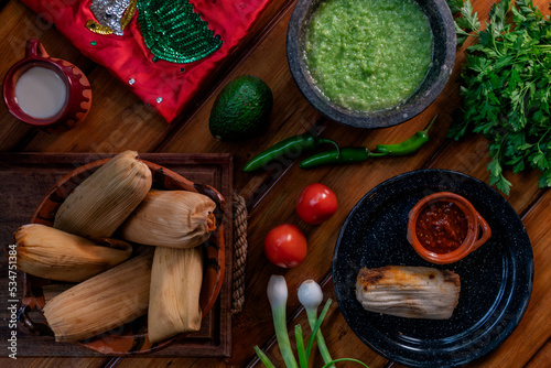 Bodegón de comida mexicana, tamales, salsa verde, salsa roja, tomate, cebolla, crema de vaca, chile, limones y queso, en platos de barro mexicano, en mesa rustica de madera y bandera de México