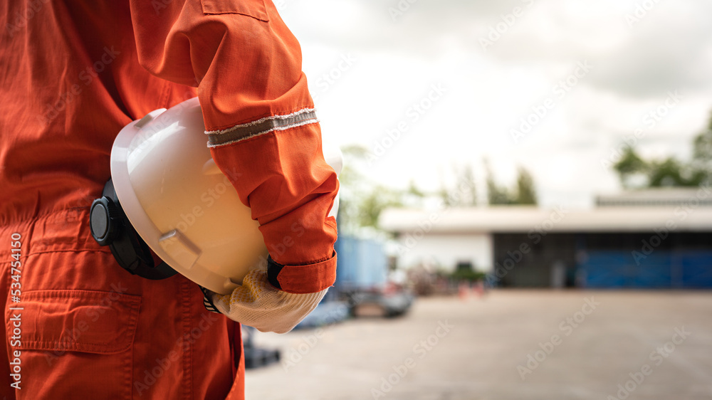 Obraz premium An operation worker in orange coverall is holding white safety helmet or hardhat with factory place as background. Safe working practice in the industrial scene photo, close-up and selective focus.