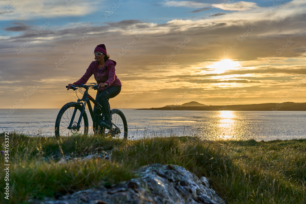 Fototapeta premium nice senior woman on mountain bike, cycling in sunset in the sand dunes of Sillerna, Grallagh, County Galway, in the western part of the Republic of Ireland