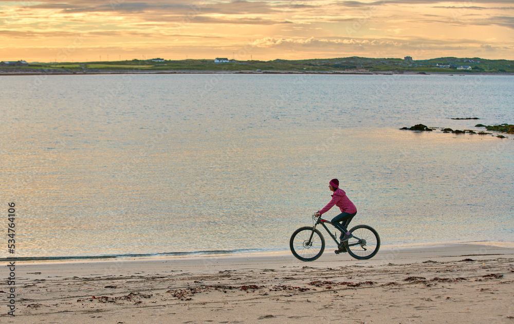 Fototapeta premium nice senior woman on mountain bike, cycling in sunset at the golden sand beach of Sillerna, Grallagh, County Galway, in the western part of the Republic of Ireland