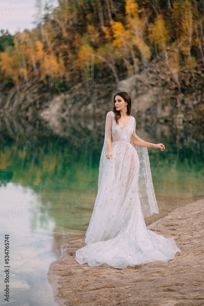 A young bride stands on the shore of a beautiful lake surrounded by an autumn forest.