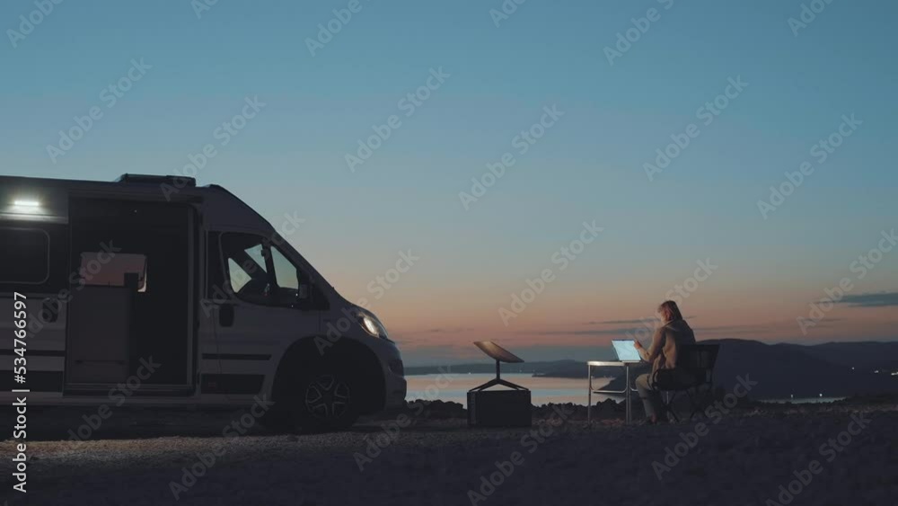 Woman working on her computer and traveling around with her camper van. She gets internet from Starlink satellite internet receiver dish. Ocean view at the sunset. Digital nomad.