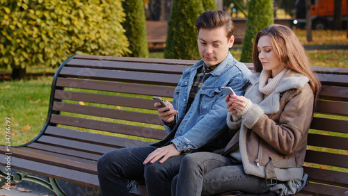 Caucasian couple two young people partners sit on bench use phone cell gadgets scroll internet web pages check social networks show images each other spend time in city park suffer of mobile addiction
