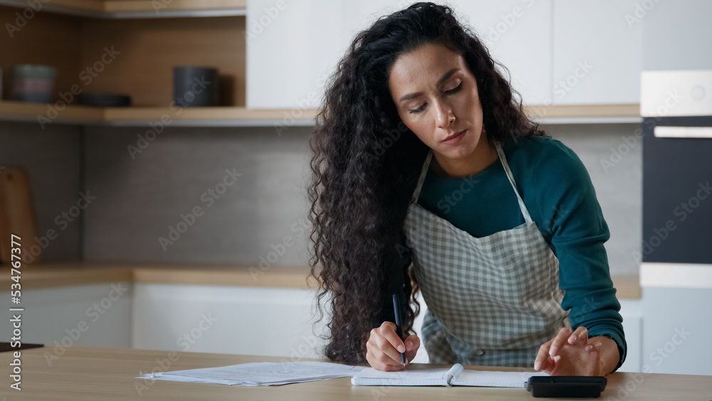 Concentrated hispanic woman wife young mom stand in modern kitchen make notes at copybook write recipe grocery list calculate domestic utilities use calculator count bills puzzled by monthly expenses Concentrated hispanic woman wife young mom stand in modern kitchen make notes at copybook write recipe grocery list calculate domestic utilities use calculator count bills puzzled by monthly expenses