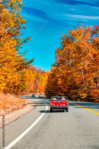 Old vintage red car driving along a road of New England in foliage season, USA