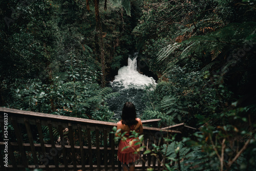 Caucasian girl standing with pink dress Calmly observing the flow of water from the waterfall among the forest trees and lush vegetation in Okere, new zealand photo