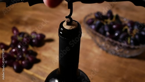 A corkscrew to open a bottle of wine in a warm atmosphere. Close-up unscrew the cork in the bottle. Hands of a man opening a bottle of red wine with a corkscrew, still life of grapes with a bottle 