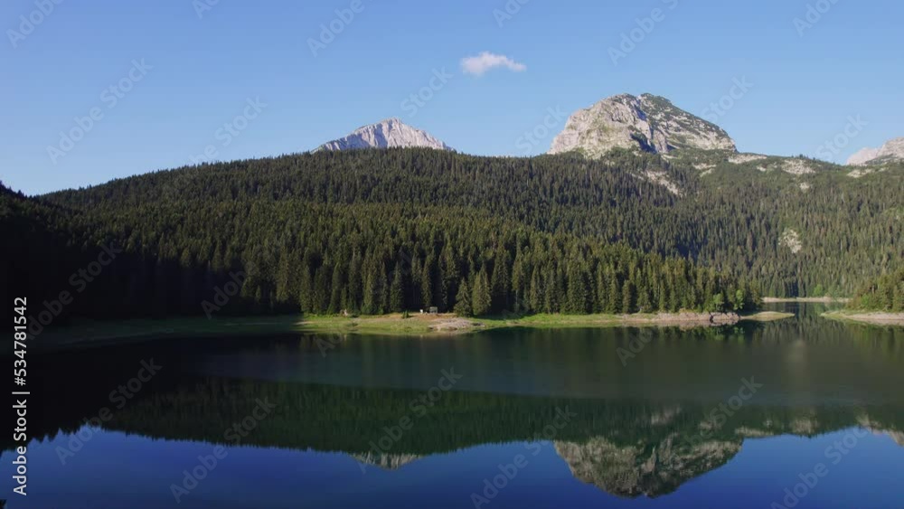 A glacial lake below a mountain peak surrounded by green coniferous forest shot from the air with a backward motion revealing the size of the lake and part of the lake's surroundings A glacial lake below a mountain peak surrounded by green coniferous forest shot from the air with a backward motion revealing the size of the lake and part of the lake's surroundings