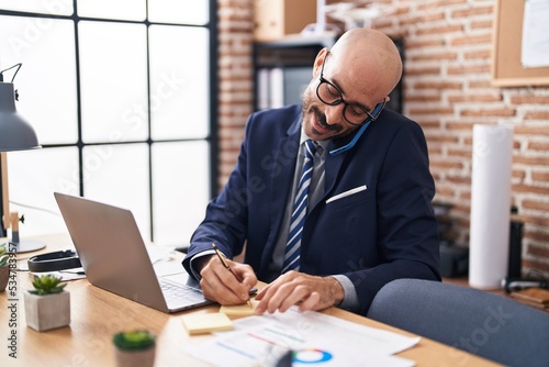 Young hispanic man business worker talking on smartphone writing on reminder paper at office