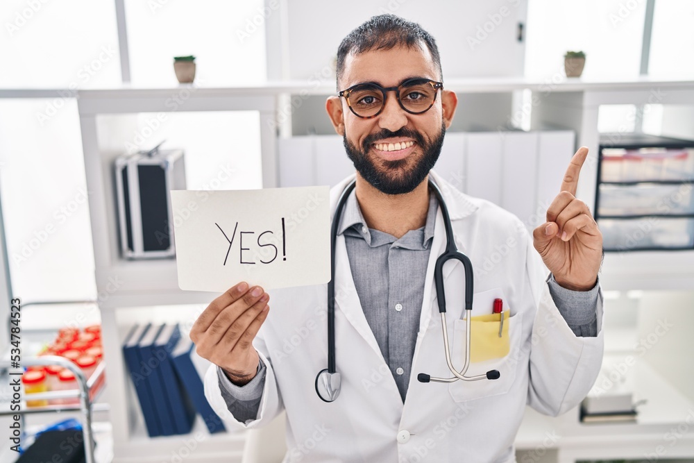 Middle east man with beard wearing doctor uniform and stethoscope holding yes banner smiling with an idea or question pointing finger with happy face, number one Middle east man with beard wearing doctor uniform and stethoscope holding yes banner smiling with an idea or question pointing finger with happy face, number one
