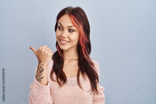 Young caucasian woman wearing pink sweater over isolated background smiling with happy face looking and pointing to the side with thumb up.