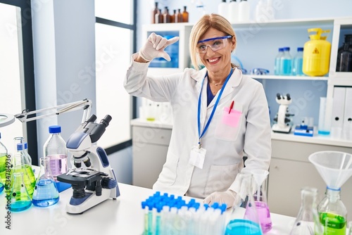 Middle age blonde woman working at scientist laboratory smiling and confident gesturing with hand doing small size sign with fingers looking and the camera. measure concept.
