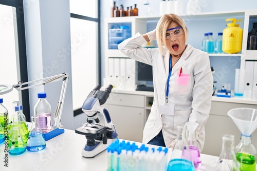 Middle age blonde woman working at scientist laboratory in shock face  looking skeptical and sarcastic  surprised with open mouth