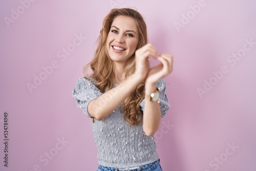 Beautiful blonde woman standing over pink background smiling in love doing heart symbol shape with hands. romantic concept.