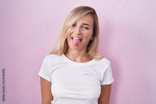 Young blonde woman standing over pink background sticking tongue out happy with funny expression. emotion concept.