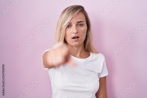 Young blonde woman standing over pink background pointing displeased and frustrated to the camera, angry and furious with you