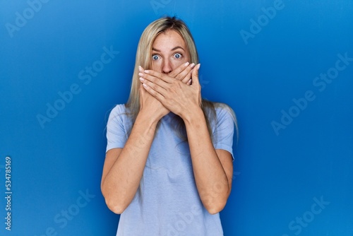 Beautiful blonde woman wearing casual t shirt over blue background shocked covering mouth with hands for mistake. secret concept.