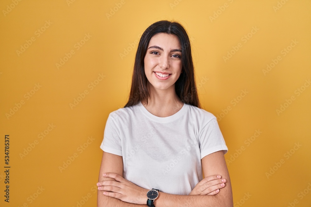 Obraz premium Young beautiful woman standing over yellow background happy face smiling with crossed arms looking at the camera. positive person.