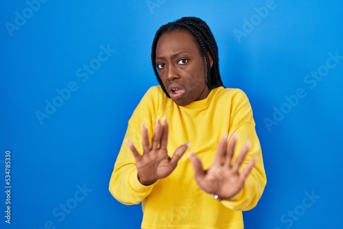 Beautiful black woman standing over blue background disgusted expression, displeased and fearful doing disgust face because aversion reaction.