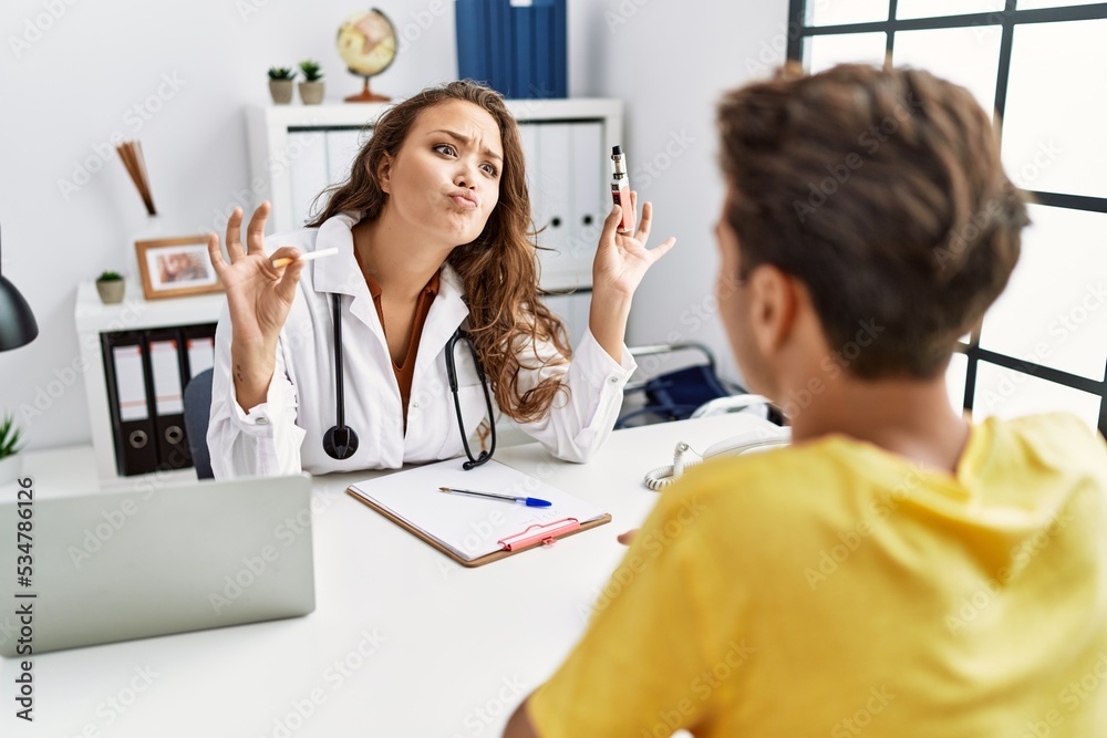 Fototapeta premium Young doctor woman showing electronic cigarette and normal cigarrete to patient looking at the camera blowing a kiss being lovely and sexy. love expression.