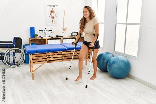 Young beautiful hispanic woman patient using crutches walking at rehab clinic