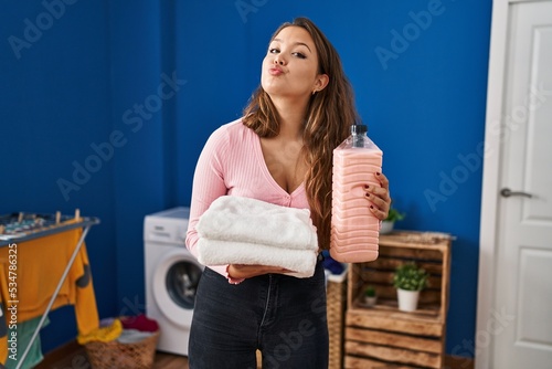 Young hispanic woman holding clean laundry and detergent bottle looking at the camera blowing a kiss being lovely and sexy. love expression.