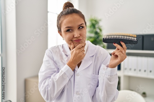 Young hispanic dentist woman holding teeth whitening samples serious face thinking about question with hand on chin, thoughtful about confusing idea