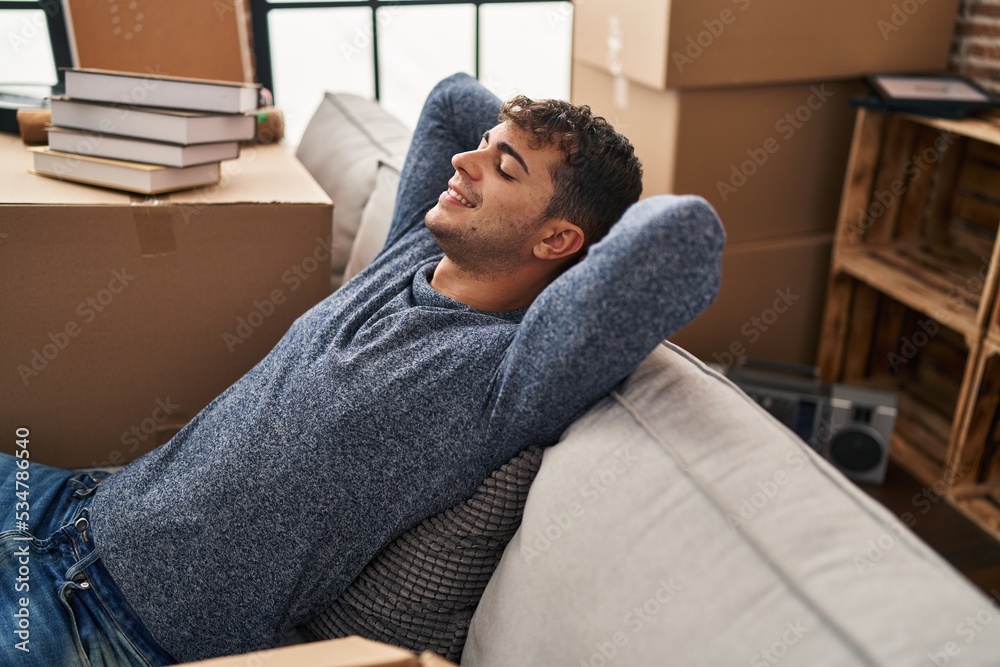 Fototapeta premium Young hispanic man relaxed with hands on head sitting on sofa at new home